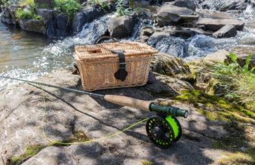 Panier à poisson en osier DELUXE (G5061) posé sur la pierre près d'un cours d'eau avec canne à moucher